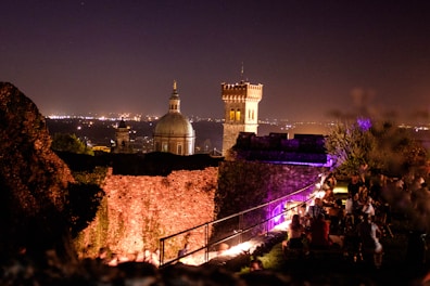 A nighttime scene featuring a historic cityscape with a prominent domed building and a tall tower. In the foreground, people are gathered around tables, enjoying an outdoor social event, illuminated by purple and yellow lights. The city lights sparkle in the background, adding a warm glow to the evening atmosphere.