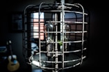 A vintage-style lamp encased in a metal cage with a visible light bulb inside. The lamp is situated in a dimly lit room with blurred background elements, including a guitar and a window with partial light coming through.