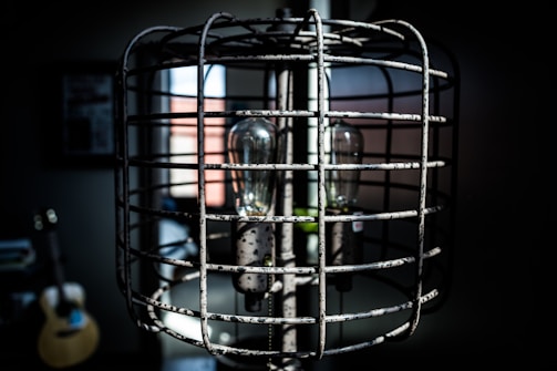 A vintage-style lamp encased in a metal cage with a visible light bulb inside. The lamp is situated in a dimly lit room with blurred background elements, including a guitar and a window with partial light coming through.