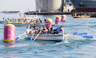 A group of rowing teams competes on clear blue waters, maneuvering colorful boats around buoy markers. Each team is focused and coordinated, dressed in matching uniforms. The background features an industrial area with large ships and harbor structures.