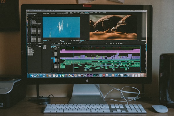 A computer monitor displays video editing software with multiple tracks of video and audio clips in a timeline. An image of aquatic life and another of a turtle are shown on the upper part of the screen. The workspace includes a keyboard, mouse, and external hard drive situated on the desk.