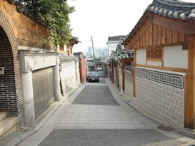 A narrow, serene alleyway lined with traditional Korean hanok houses features intricate wooden architecture and tiled roofs. The cobblestone path gently slopes downwards, with a parked car visible in the distance. Lush greenery from an overhanging tree adds a natural element to the setting.