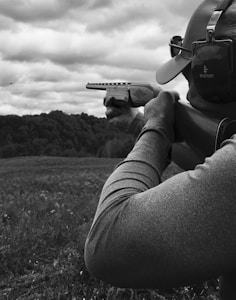 A person wearing a cap and protective earmuffs is aiming a shotgun in an outdoor setting. The landscape features an open field and a wooded hillside in the background under a cloudy sky.