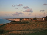 A coastal landscape featuring white cliffs extending along the horizon, with a grassy field in the foreground. Several wind turbines are positioned near a few scattered buildings, and the sky is warm with scattered clouds during what appears to be sunset.