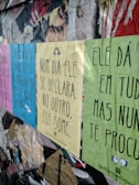 Posters with inspirational and introspective quotes are pasted on a worn and textured urban wall. The colors of the posters are pastel pink, blue, yellow, and green. The wall shows signs of decay with peeling paint and exposed layers.