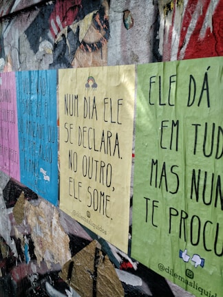 Posters with inspirational and introspective quotes are pasted on a worn and textured urban wall. The colors of the posters are pastel pink, blue, yellow, and green. The wall shows signs of decay with peeling paint and exposed layers.