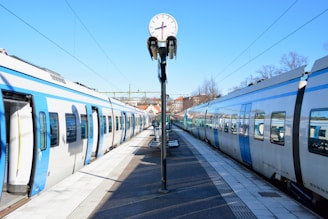 A railway station platform with two modern trains facing each other on either side. A clock is centrally positioned above the platform, showing the time. The sky is clear and blue, and there are trees and buildings visible in the background.