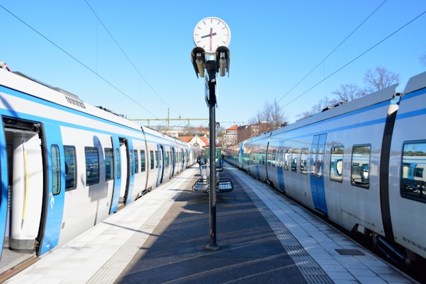A railway station platform with two modern trains facing each other on either side. A clock is centrally positioned above the platform, showing the time. The sky is clear and blue, and there are trees and buildings visible in the background.