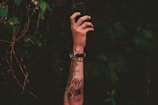 A tattooed arm extends upward holding a smartphone against a backdrop of dark green foliage. The tattoo includes an intricate design with a watch on the wrist, capturing a moment in nature.