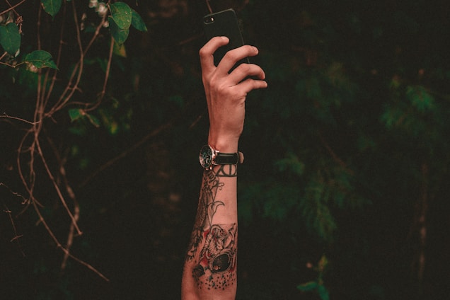 A tattooed arm extends upward holding a smartphone against a backdrop of dark green foliage. The tattoo includes an intricate design with a watch on the wrist, capturing a moment in nature.