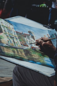 An artist is painting a picturesque scene featuring a row of old buildings along a canal in a possibly European town. The painting is vibrant with the use of various colors and intricate details of the structures and their reflections in the water.