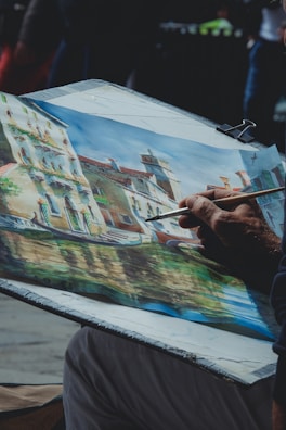 An artist is painting a picturesque scene featuring a row of old buildings along a canal in a possibly European town. The painting is vibrant with the use of various colors and intricate details of the structures and their reflections in the water.