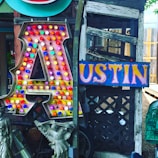 A large, illuminated letter 'A' covered with colorful bulbs stands next to a vibrant sign spelling 'USTIN'. The background features weathered wood and lattice, while statues and various decorative items are placed nearby.