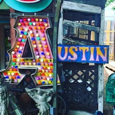 A large, illuminated letter 'A' covered with colorful bulbs stands next to a vibrant sign spelling 'USTIN'. The background features weathered wood and lattice, while statues and various decorative items are placed nearby.
