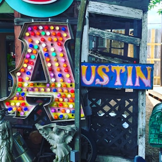 A large, illuminated letter 'A' covered with colorful bulbs stands next to a vibrant sign spelling 'USTIN'. The background features weathered wood and lattice, while statues and various decorative items are placed nearby.