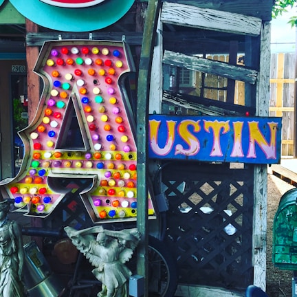 A large, illuminated letter 'A' covered with colorful bulbs stands next to a vibrant sign spelling 'USTIN'. The background features weathered wood and lattice, while statues and various decorative items are placed nearby.