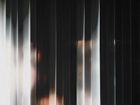 Vertical blinds create an abstract pattern, with soft light and shadow interplay, producing a gradient effect across the slats. The overall atmosphere is mysterious with hints of light peeking through gaps.
