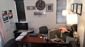 An office setting with a large wooden desk at its center. On the desk are various items including a laptop, a desktop computer, a computer monitor, a vase of pink flowers, and a printer. The walls are decorated with multiple framed pictures and a large clock. There are posters on one wall, and two windows with blinds partially open. A lamp illuminates the workspace.