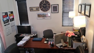 An office setting with a large wooden desk at its center. On the desk are various items including a laptop, a desktop computer, a computer monitor, a vase of pink flowers, and a printer. The walls are decorated with multiple framed pictures and a large clock. There are posters on one wall, and two windows with blinds partially open. A lamp illuminates the workspace.