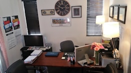 An office setting with a large wooden desk at its center. On the desk are various items including a laptop, a desktop computer, a computer monitor, a vase of pink flowers, and a printer. The walls are decorated with multiple framed pictures and a large clock. There are posters on one wall, and two windows with blinds partially open. A lamp illuminates the workspace.