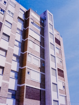 A tall urban apartment building with a mix of beige and brown brickwork. The structure features multiple balconies and white window frames, with many shutters closed. The facade displays a pattern of alternating materials, including some concrete sections. The sky in the background is a clear and bright blue.