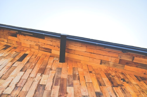 A close-up view of a wooden roof under a clear blue sky. The wooden planks are arranged in a layered pattern creating a warm, rustic aesthetic. The deep angle emphasizes the geometric structure, while a metal gutter runs along the edge.