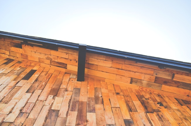A close-up view of a wooden roof under a clear blue sky. The wooden planks are arranged in a layered pattern creating a warm, rustic aesthetic. The deep angle emphasizes the geometric structure, while a metal gutter runs along the edge.