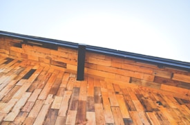A close-up view of a wooden roof under a clear blue sky. The wooden planks are arranged in a layered pattern creating a warm, rustic aesthetic. The deep angle emphasizes the geometric structure, while a metal gutter runs along the edge.