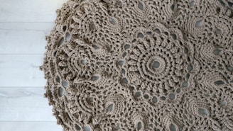 An intricately crocheted round doily with a detailed floral pattern, displayed on a light wooden floor. The texture of the yarn is coarse, giving it an artisanal look.