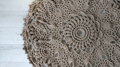 An intricately crocheted round doily with a detailed floral pattern, displayed on a light wooden floor. The texture of the yarn is coarse, giving it an artisanal look.