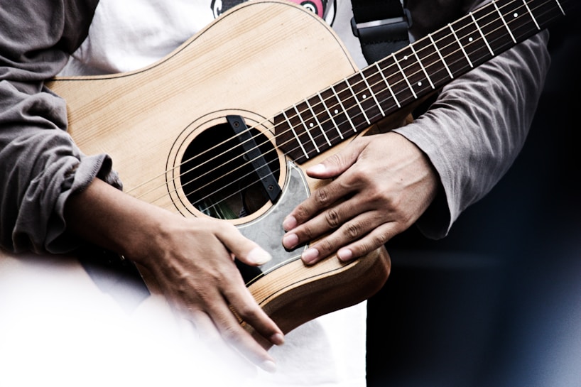 A close-up of a person playing an acoustic guitar, focusing on their hands and the wooden body of the instrument. The person's sleeves are visible, and they appear to be strumming or preparing to play.