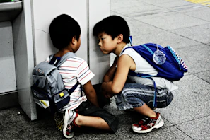 Two young boys are sitting on the floor, facing each other, with serious expressions. They both have backpacks, indicating they might be in a school or travel setting. The setting appears to be an urban environment with a tiled floor and a wall providing a corner for them to sit against.