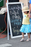 A chalkboard sign advertises cupcakes that are baked fresh, gluten-free, and vegan. The sign is leaning against a table or booth at what appears to be an outdoor market. A child in a bright blue skirt and a yellow shirt is walking by, holding hands with an adult. The child is wearing a hat that partially obscures their face.