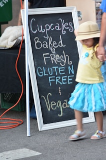 A chalkboard sign advertises cupcakes that are baked fresh, gluten-free, and vegan. The sign is leaning against a table or booth at what appears to be an outdoor market. A child in a bright blue skirt and a yellow shirt is walking by, holding hands with an adult. The child is wearing a hat that partially obscures their face.