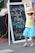 A chalkboard sign advertises cupcakes that are baked fresh, gluten-free, and vegan. The sign is leaning against a table or booth at what appears to be an outdoor market. A child in a bright blue skirt and a yellow shirt is walking by, holding hands with an adult. The child is wearing a hat that partially obscures their face.