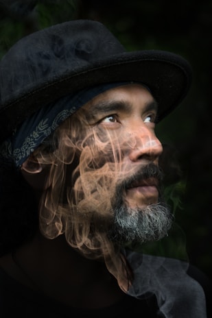 A person with a contemplative expression is looking upward, wearing a dark hat and a patterned bandana. A thick, swirling smoke effect overlays the image, adding a mysterious and surreal quality.