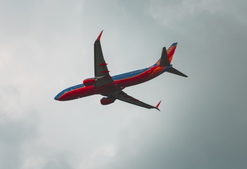 A brightly colored commercial airplane with red, blue, and orange hues flies through a cloudy sky. The aircraft is in motion, appearing to ascend or maintain elevation, with its wings and tail tips prominently visible.
