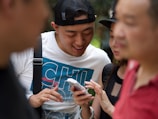 A group of people are gathered closely together, focusing on a smartphone held by one person. The central figure is smiling and appears to be sharing something amusing or interesting. The setting seems to be outdoors with blurred greenery in the background.