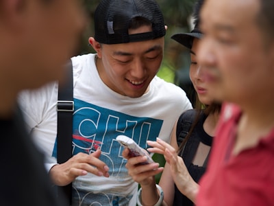 A group of people are gathered closely together, focusing on a smartphone held by one person. The central figure is smiling and appears to be sharing something amusing or interesting. The setting seems to be outdoors with blurred greenery in the background.