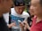 A group of people are gathered closely together, focusing on a smartphone held by one person. The central figure is smiling and appears to be sharing something amusing or interesting. The setting seems to be outdoors with blurred greenery in the background.