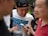 A group of people are gathered closely together, focusing on a smartphone held by one person. The central figure is smiling and appears to be sharing something amusing or interesting. The setting seems to be outdoors with blurred greenery in the background.