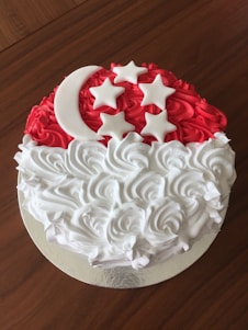 A circular cake decorated with red and white swirled frosting. The top half features a red background with a white crescent moon and five stars made of icing, while the bottom half is adorned with white swirled frosting.