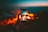 A hand holds a smartphone capturing a photo of a campfire in a dark outdoor setting. The background is filled with warm tones from the fire, contrasting against the deep blue twilight sky.