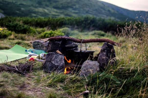 A campfire is set up with a makeshift cooking area, consisting of pots hanging over a fire on a wooden stick supported by rocks. In the foreground, there are grass and some branches scattered around. Nearby, a sleeping mat and picnic items, including a plastic bottle, are visible. The background displays lush greenery with a rolling hill under a cloudy sky.
