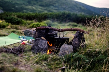 A campfire is set up with a makeshift cooking area, consisting of pots hanging over a fire on a wooden stick supported by rocks. In the foreground, there are grass and some branches scattered around. Nearby, a sleeping mat and picnic items, including a plastic bottle, are visible. The background displays lush greenery with a rolling hill under a cloudy sky.