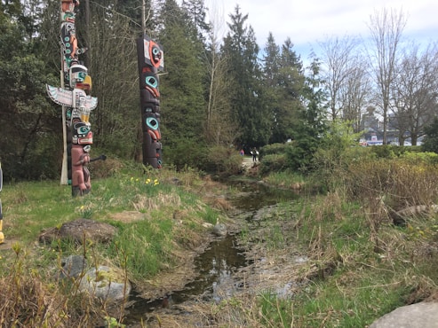 Artistic totem poles stand among lush greenery and trees near a small creek, with a few people in the distance walking along a path. The scene is serene and reflects both nature and cultural art.