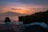 A stunning sunset over an ocean cliff, with vibrant hues of orange and pink in the sky. A lone figure is captured mid-jump off the edge of the cliff into the sea below. There are dark, rugged cliffs to the side, and the waves crash against the rocks, highlighted by the fading daylight.