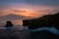 A stunning sunset over an ocean cliff, with vibrant hues of orange and pink in the sky. A lone figure is captured mid-jump off the edge of the cliff into the sea below. There are dark, rugged cliffs to the side, and the waves crash against the rocks, highlighted by the fading daylight.
