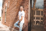 A person in a white t-shirt and ripped jeans is leaning against a brick wall. The scene is urban, with the reflection of a tree and buildings visible in a large window. The image highlights casual fashion and has a relaxed atmosphere.