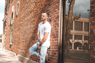 A person in a white t-shirt and ripped jeans is leaning against a brick wall. The scene is urban, with the reflection of a tree and buildings visible in a large window. The image highlights casual fashion and has a relaxed atmosphere.